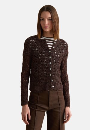 Mujer con un cárdigan marrón oscuro de punto abotonado sobre una camisa a rayas y pantalones marrones a juego, de pie frente a un fondo liso.