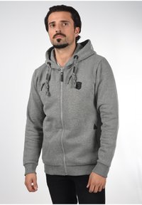 Sivi zip-up dukserica s kapuljačom na vezanje, dugim rukavima i malim logotipom. Riblje manžete i rub, teksturirani materijal.