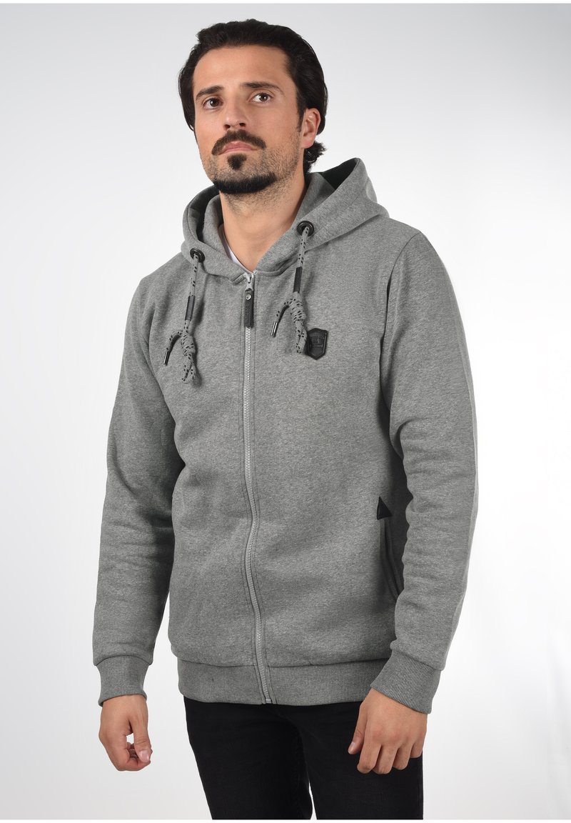 Sivi zip-up dukserica s kapuljačom na vezanje, dugim rukavima i malim logotipom. Riblje manžete i rub, teksturirani materijal.