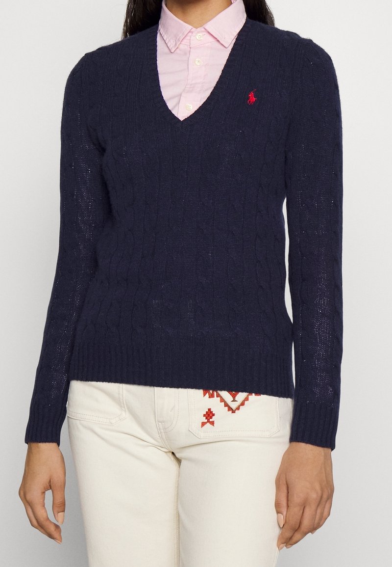 Femme portant un pull en tricot torsadé bleu marine à col en V, par-dessus une chemise à col rose clair et un pantalon blanc avec une broderie rouge.