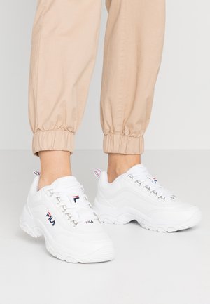 Hvide FILA chunky sneakers båret med beige opsmøgede bukser, der viser front- og sidevisning på en ensartet lys baggrund.