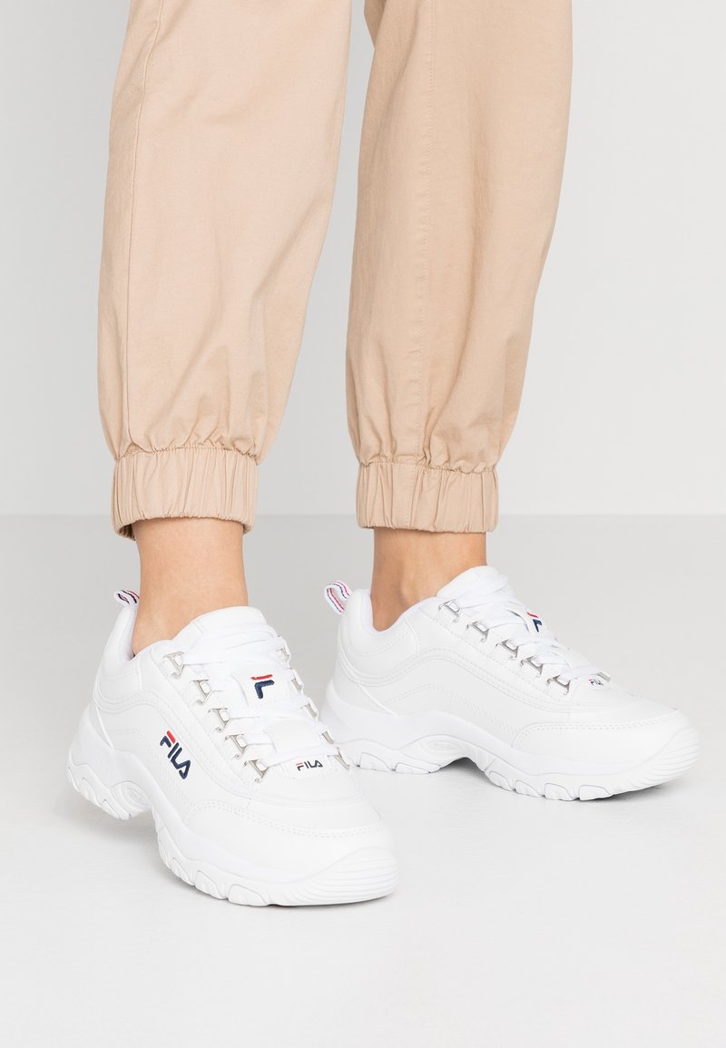 Baskets épaisses blanches FILA portées avec un pantalon beige retroussé, vues de face et de côté sur un fond clair uni.