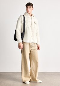 Kremfarget fleecejakke med store lommer, kombinert med beige vide bukser. Tilbehør inkluderer en svart skulderveske og hvite sko.