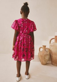 Pink paljetkjole med korte pufærmer, blomsterprint og knaplukning bagpå. Hvide sko fuldender outfittet.