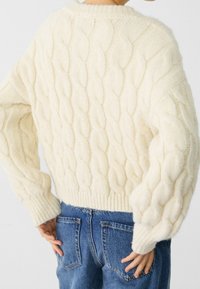 Dik crèmekleurig kabelgebreide trui met een ronde halslijn en lange, oversized mouwen; geribbelde zoom; gedragen met blauwe spijkerbroek.