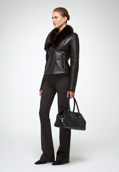 Donna in giacca di pelle nera con colletto in pelliccia, pantaloni grigi scuri su misura, scarpe nere a punta, che tiene una borsa di pelle nera, in piedi di lato.