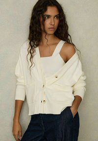 Cardigan bianco lavorato a maglia con grandi bottoni frontali e due tasche, indossato sopra un top di colore simile, abbinato a pantaloni in denim scuri.