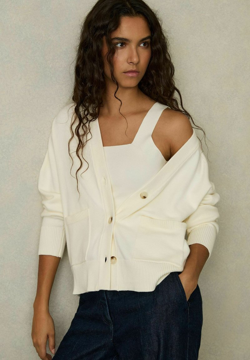 Cardigan bianco lavorato a maglia con grandi bottoni frontali e due tasche, indossato sopra un top di colore simile, abbinato a pantaloni in denim scuri.