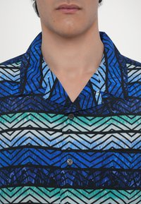 Cămașă cu mânecă scurtă, cu nasturi, prezentând un model geometric în nuanțe de albastru și teal, cu accente negre și guler. Textură de material fin.