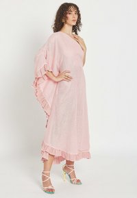 Mujer con cabello rizado posa con un vestido suelto color rosa claro con bordes fruncidos y sandalias de tacón multicolores sobre un fondo blanco.