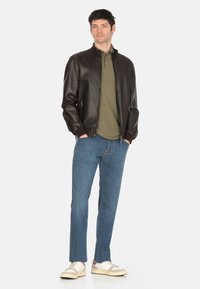 Giubbotto bomber in pelle marrone con polsini a costine, polo verde oliva, jeans azzurri chiari e sneaker beige. Texture liscia e design aderente.