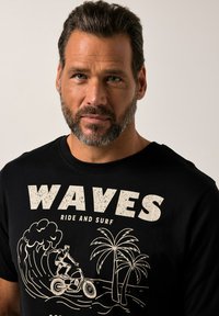 Zwarte T-shirt met een grafische print met de tekst "WAVES RIDE AND SURF," een surfer op een fiets, golven en palm bomen in crèmekleur.