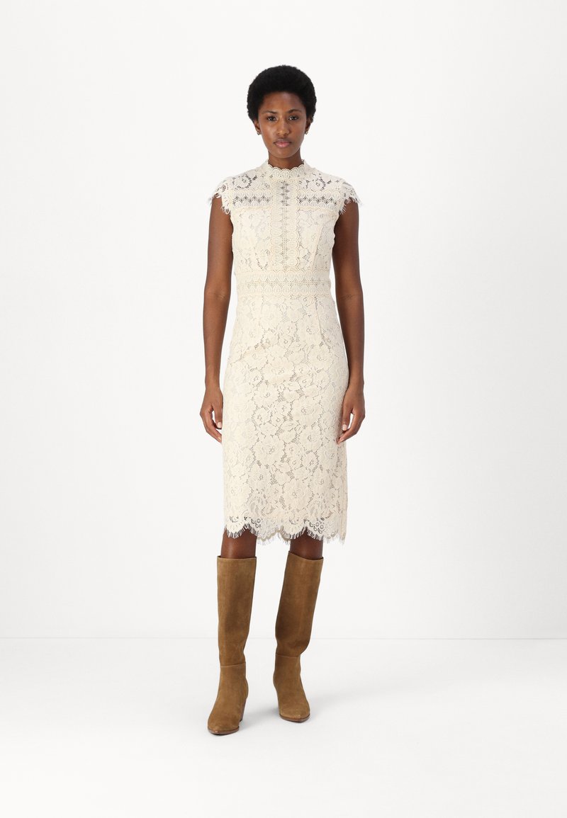 Robe en dentelle blanc cassé avec un col haut, sans manches et un ourlet en forme de vagues, associée à des bottes en suède marron jusqu'aux genoux.