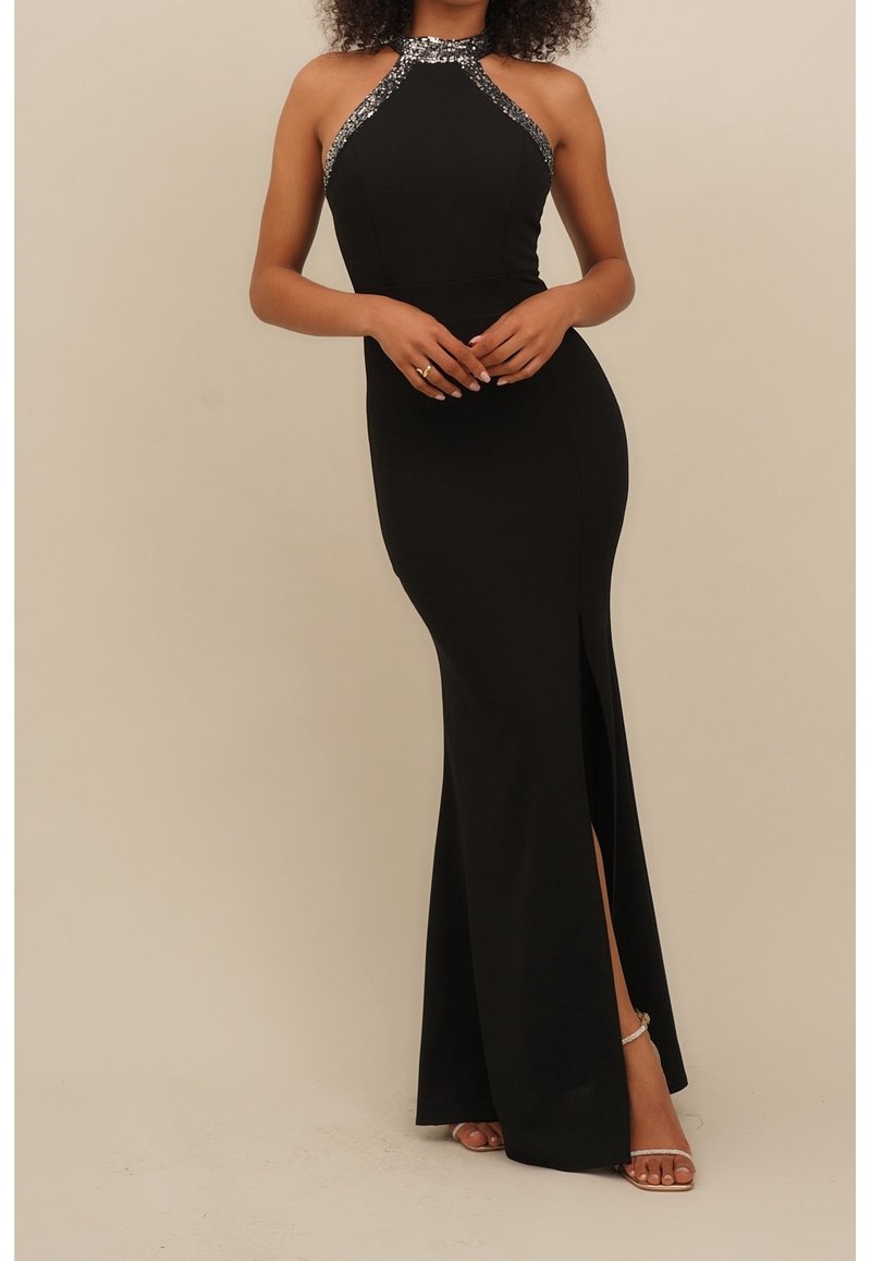 Femme portant une robe noire sans manches avec un bord en sequins, une fente sur le côté, chaussée de talons ouverts, se tenant les mains croisées à la taille.