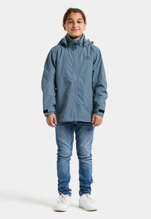 Veekindel sinine jope kapuutsiga, kontrastse halli voodriga, ees tõmblukuga ja mustade detailidega. Kandmisel sinised teksad ja valged jalanõud.