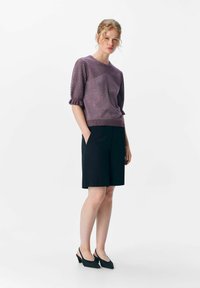 Fioletowy sweter z dzianiny z teksturowanymi krótkimi rękawami, zestawiony z czarnymi dopasowanymi szortami. Modelka stoi w czarnych sandałach na obcasie na jednolitym tle.