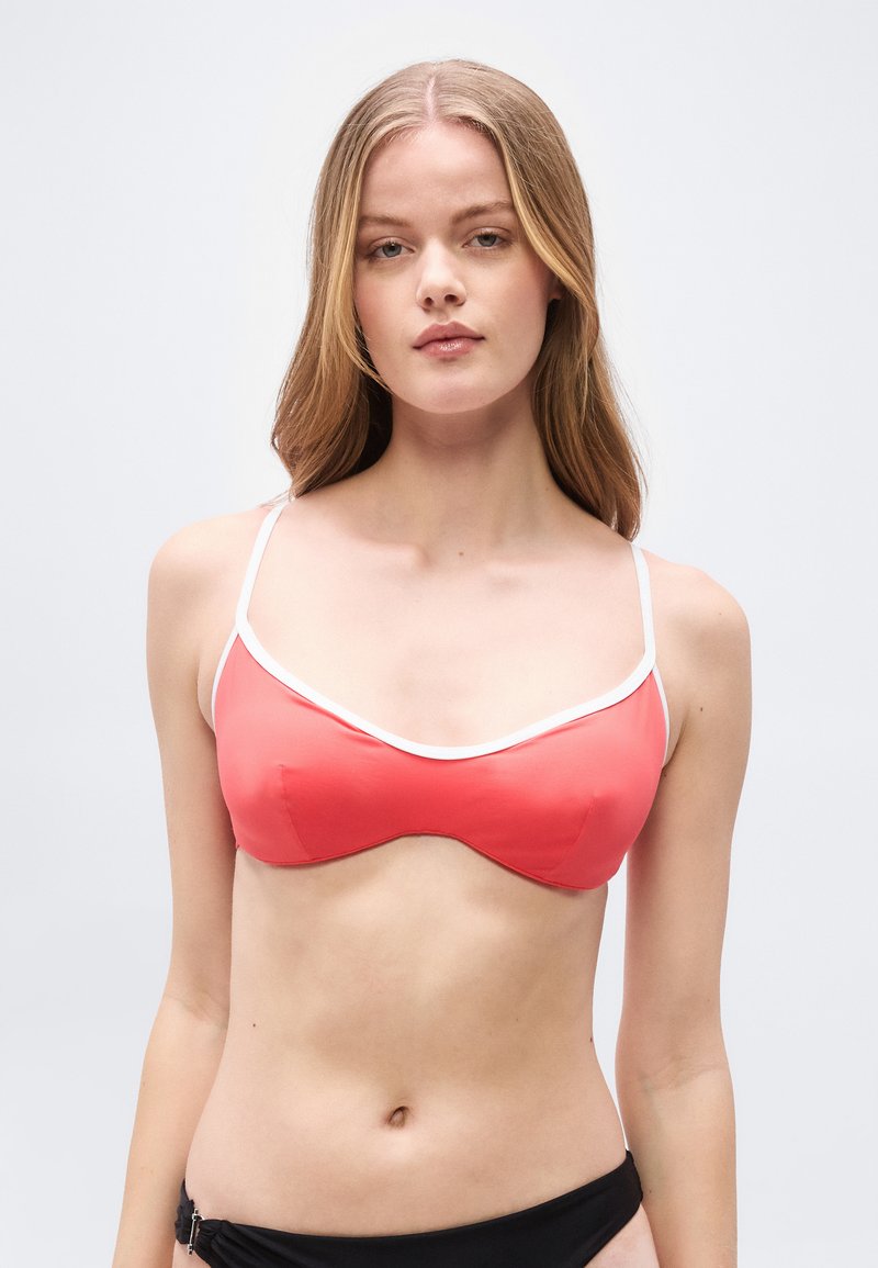 Νεαρή γυναίκα με μακριά ξανθά μαλλιά, φοράει τοπ μπικίνι κοραλί ροζ με λευκή διακόσμηση και μαύρο κάτω μέρος μπικίνι, κοιτάζοντας μπροστά σε απλό φόντο.