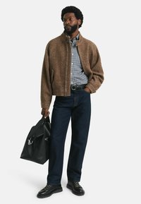 Chaqueta de espiga marrón sobre una camisa a cuadros azul y blanca, combinada con jeans oscuros y zapatos de cordones negros, sosteniendo una bolsa de mano negra.