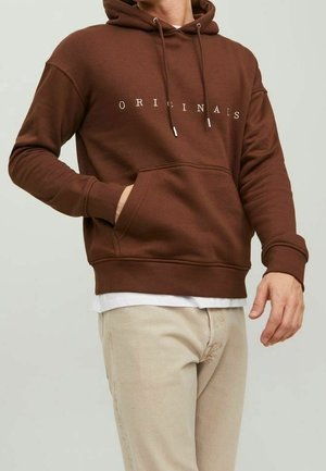 Ruskea puuvillainen huppari kengurutaskulla, kiristysnauhoilla ja valkoisella "ORIGINALS"-tekstillä, kerrospukeutunut valkoisen paidan ja beigen housujen kanssa.