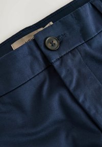 Gros plan de la ceinture d'un pantalon bleu marine avec un bouton noir attaché et une étiquette de vêtement beige partiellement visible à l'intérieur.