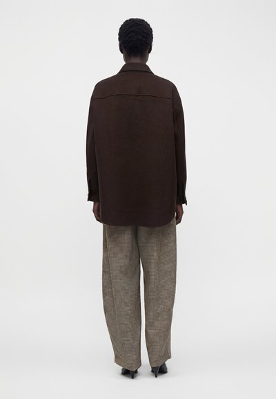 Personne vue de dos portant une surchemise marron foncé, un pantalon large à carreaux et des chaussures à talons noires sur un fond clair uni.