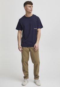 Marineblå bomulds T-shirt med korte ærmer, prydet med et lille hvidt logo på venstre side. Sammen med beige bukser og hvide sko.