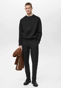Felpa nera con polsini a coste e colletto a girocollo, abbinata a pantaloni coordinati. Una giacca marrone appoggiata sul braccio, scarpe nere completano il look.
