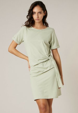 Vrouw met donker golvend haar, gekleed in een lichtgroene jurk met korte mouwen en een knoopdetail aan de zijkant, staand met één hand op de heup.