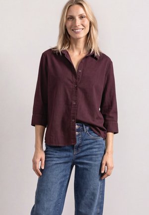 Lyshåret kvinde smiler iført en burgunder knap-op skjorte med trekvartærmer og blå jeans mod en ensartet lys baggrund.