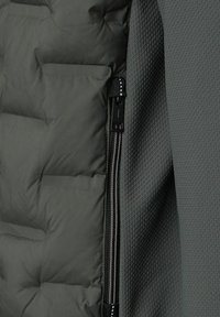 Närbild av en mörkgrå pufferjacka som visar en svart dragkedja mellan slät vadderad tyg och texturerat nätmaterial.