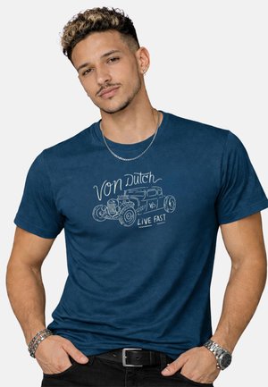 Jeune homme aux cheveux bouclés portant un t-shirt bleu avec un dessin d'une voiture vintage et le texte "Von Dutch Live Fast" en blanc.