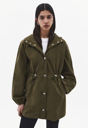 Donna con capelli lunghi e scuri che indossa una giacca con cappuccio verde oliva con bottoni frontali e coulisse in vita, in piedi su uno sfondo bianco.