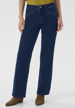 Jean en denim bleu foncé avec une coupe droite, présentant des poches avant et des détails de couture visibles. Associé à des bottines brunes.
