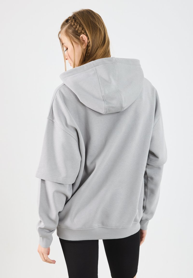 Sudadera con capucha gris, hecha de tela suave, con un diseño oversized, hombros caídos y puños acanalados. Sin patrones ni adornos visibles.