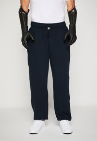 Mørkeblå joggebukser med snøring i midjen, laget av mykt stoff. Sammen med hvite joggesko. Robuste svarte hansker på hendene.