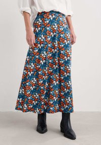 Gėlių maksi sijonas su mėlynu fonu, oranžinėmis, baltomis ir geltonomis gėlių raštais, pagamintas iš lengvos medžiagos, derinamas su juodomis botinėmis.