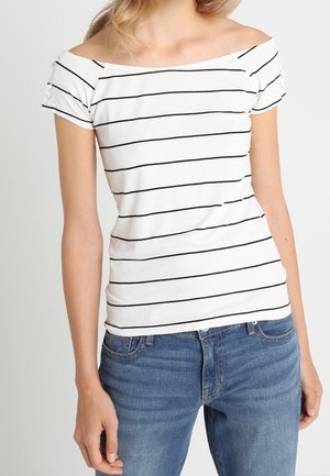 Vrouw draagt een witte off-shoulder top met zwarte horizontale strepen en blauwe spijkerbroek, staand tegen een neutrale achtergrond.