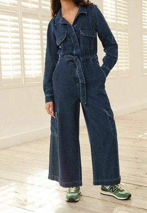Combinaison en denim bleu foncé, dotée d'un col, de manches longues, de deux poches poitrine, de jambes larges et d'une taille ajustée par un lien. Soulignée par des coutures blanches.