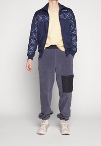 Persona con chaqueta de chándal de Adidas de patrón azul marino, camiseta beige, pantalones de chándal grises con bolsillo negro, zapatillas blancas, de pie contra un fondo liso.