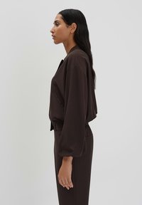 Brown cropped jacket with long, voluminous sleeves and side pockets. Smooth fabric drapes gently. Model wearing matching pants stands side profile.