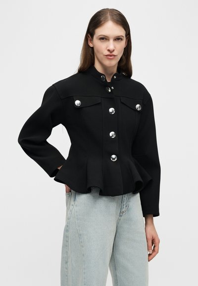 Γυναίκα με ίσια καστανά μαλλιά, φοράει μαύρο σακάκι με ασημί κουμπιά και ανοιχτό μπλε τζιν, χέρια στις τσέπες, ουδέτερο γκρι φόντο.