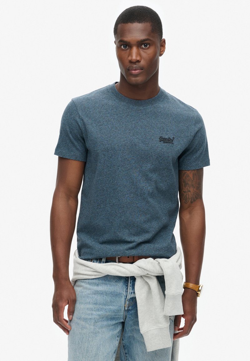 T-shirt in cotone blu con texture, colletto girocollo e maniche corte, con un piccolo logo nero sul petto. Abbinato a jeans chiari.