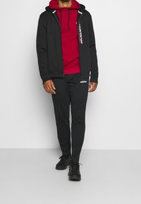 Schwarze Zip-Hoodie über einem roten Sweatshirt, kombiniert mit schwarzen Hosen mit Adidas-Logo; der Stoff ist glatt und hat eine lockere Passform.