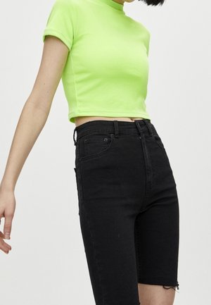 Persoon draagt een felgroen cropped t-shirt en een zwarte high-waisted denim short tegen een effen lichte achtergrond.