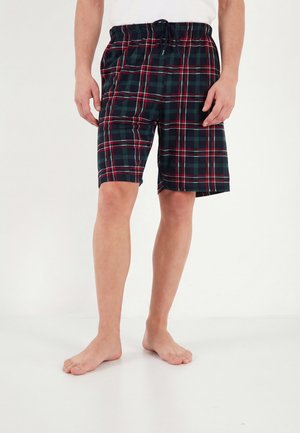 Mand iført mørkegrønne, røde og hvide ternede shorts med en sort snøre, står barfodet på en hvid gulv mod en hvid baggrund.