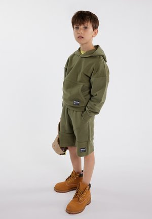 Sweat à capuche en polaire vert olive avec poche frontale, associé à un short assorti. Bottes en cuir marron avec des lacets jaunes et des logos sur les vêtements.