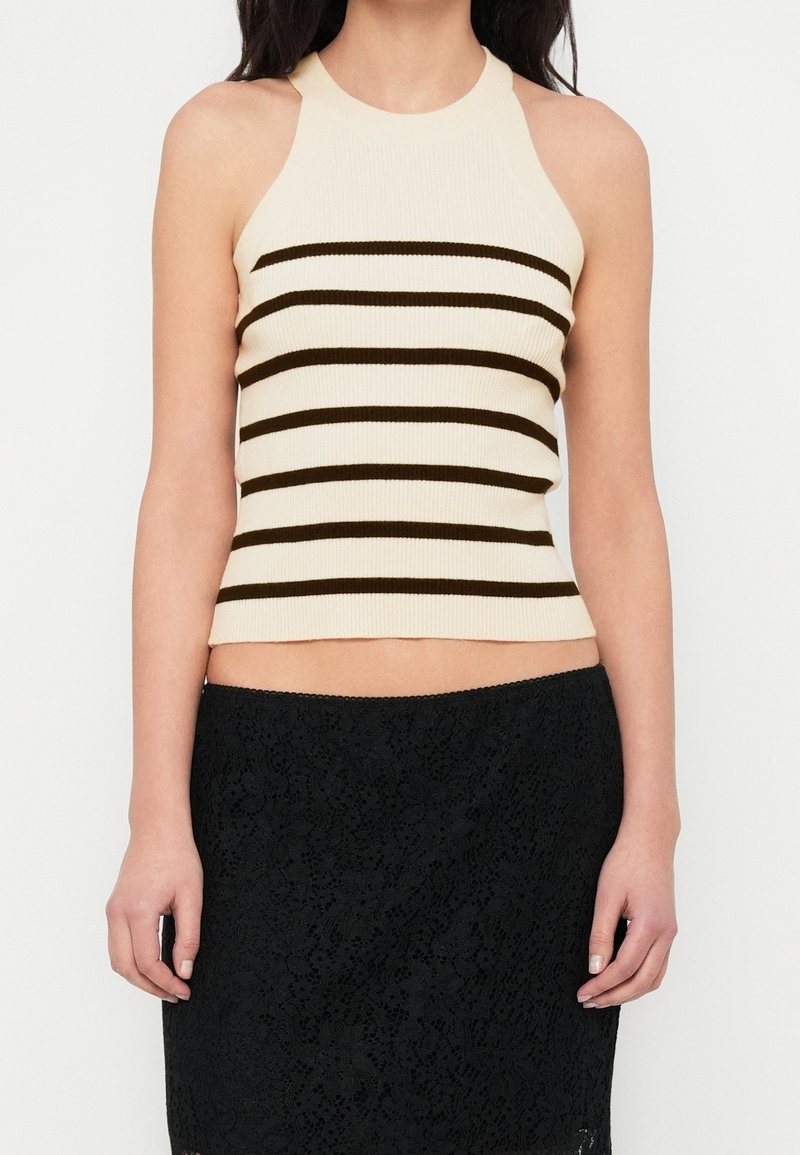 Femme portant un haut sans manches beige en maille côtelée avec des rayures horizontales noires et une jupe en dentelle noire, debout devant un fond blanc.
