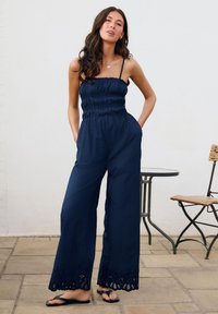 Mono azul marino con cuerpo fruncido, piernas anchas y ribete de encaje en el dobladillo. Combinado con sandalias negras tipo chancla, sobre un fondo claro.