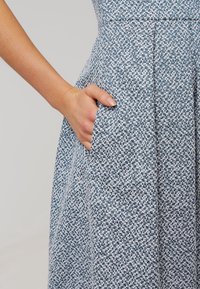 La mano di una donna è appoggiata all'interno della tasca di una gonna o di un vestito plissettato blu e bianco con texture.