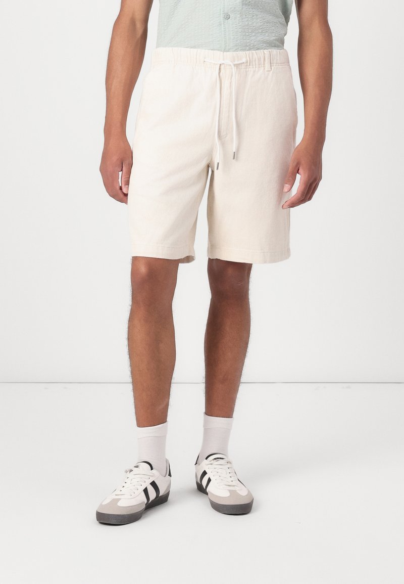 Shorts beige clair avec une taille élastique et un cordon de serrage ; fabriqués en tissu doux, atteignant mi-cuisse, avec des poches latérales.